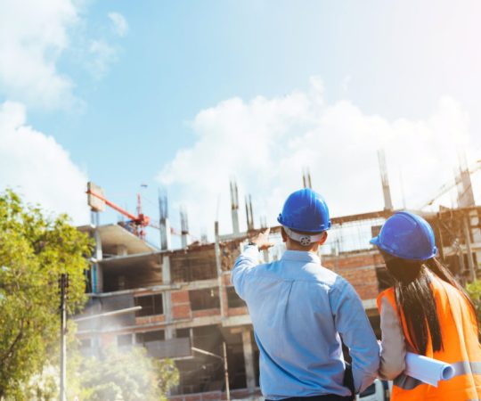 Asian man civil engineer and woman architect wearing blue safety helmet meeting at contruction site.